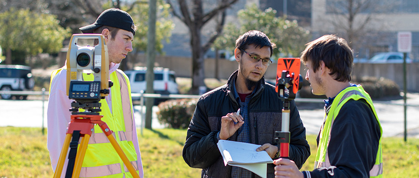 two undergraduate students work with surveying equipment outside, led by a graduate student.